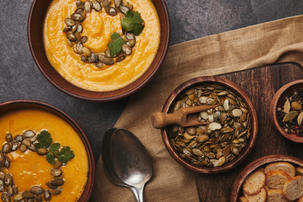 top view of bowls with pumpkin soup, spoons, rusks and pumpkin seeds on wooden table